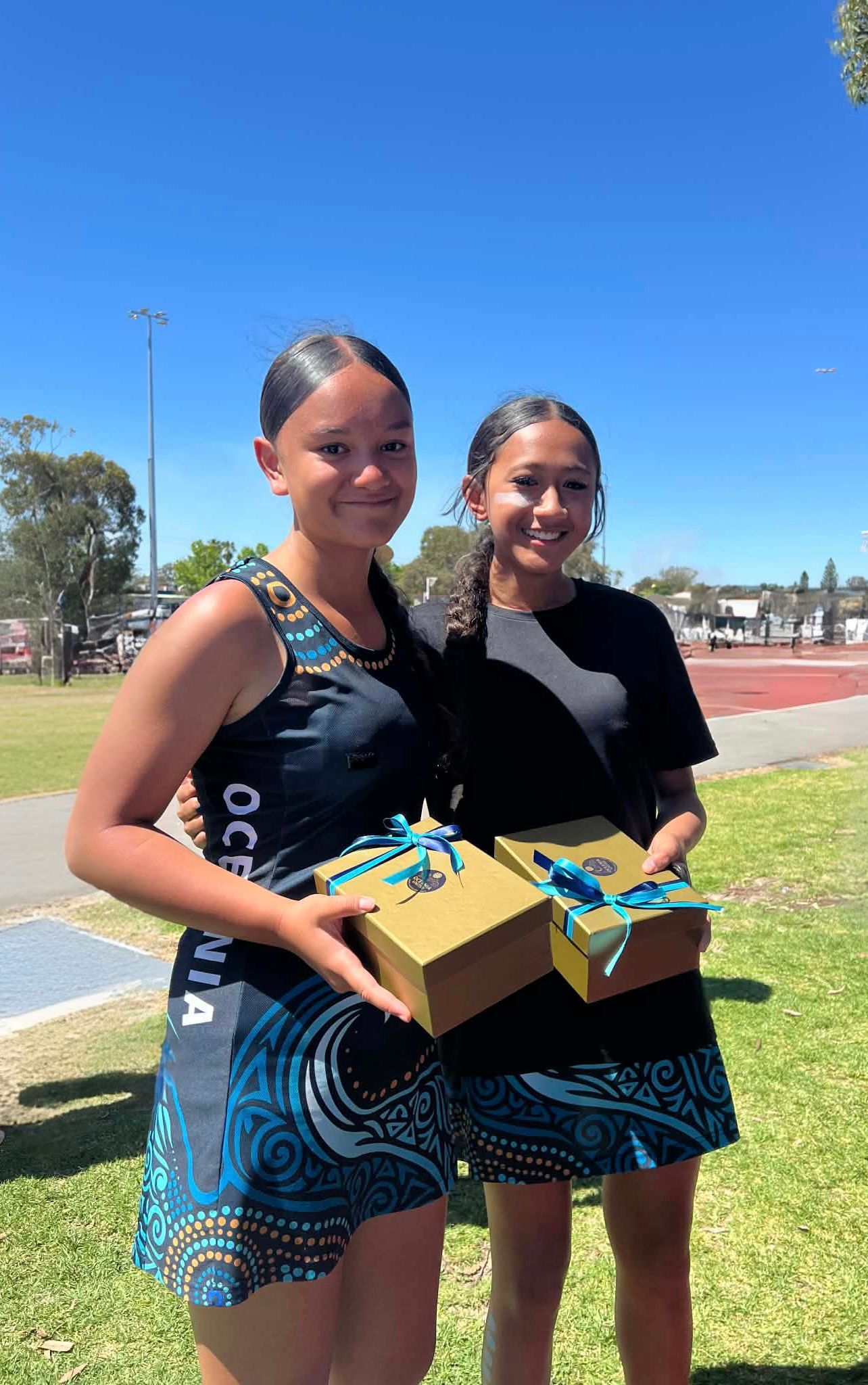 Two young women in matching athletic outfits, holding gift boxes outdoors on a sunny day.