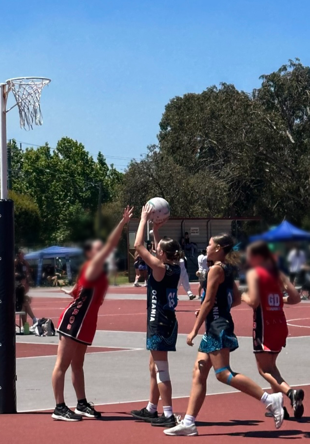 Netball players on a court, one shooting toward the net, others defending and moving. Sunny outdoor setting.