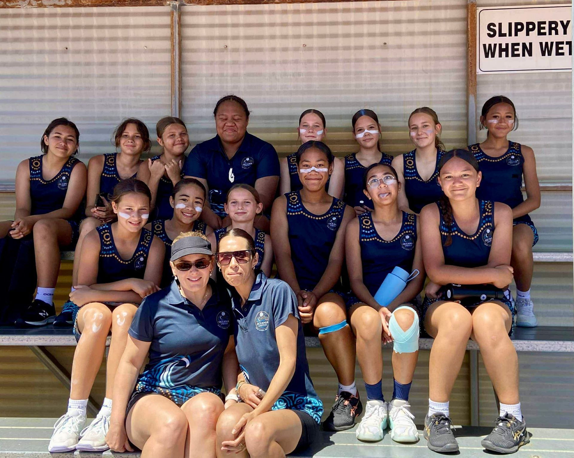 Netball team posing, navy uniforms, sitting on bench under a 