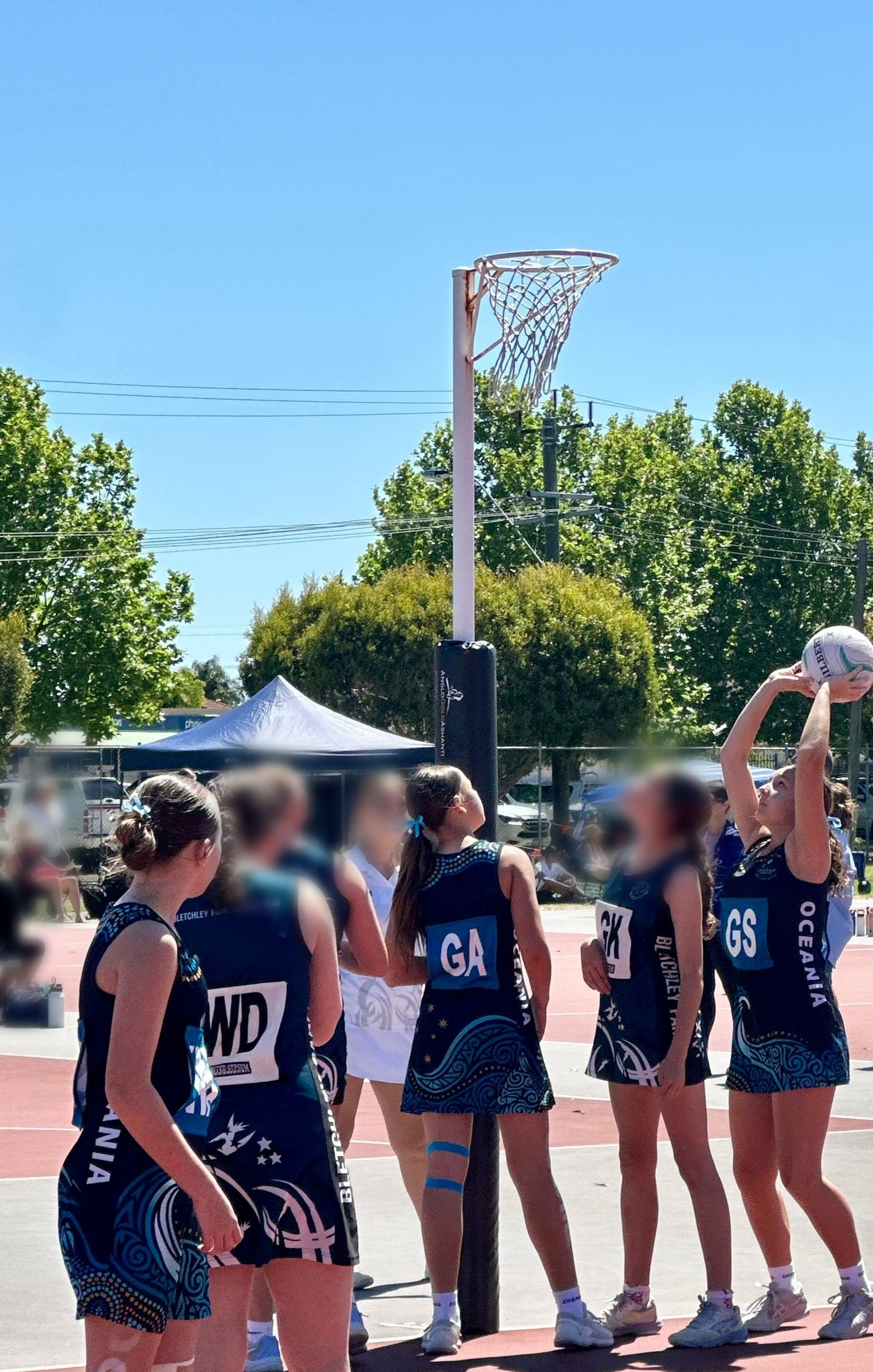 Netball players in blue uniforms; one shoots towards the net. Sunny day at an outdoor court.