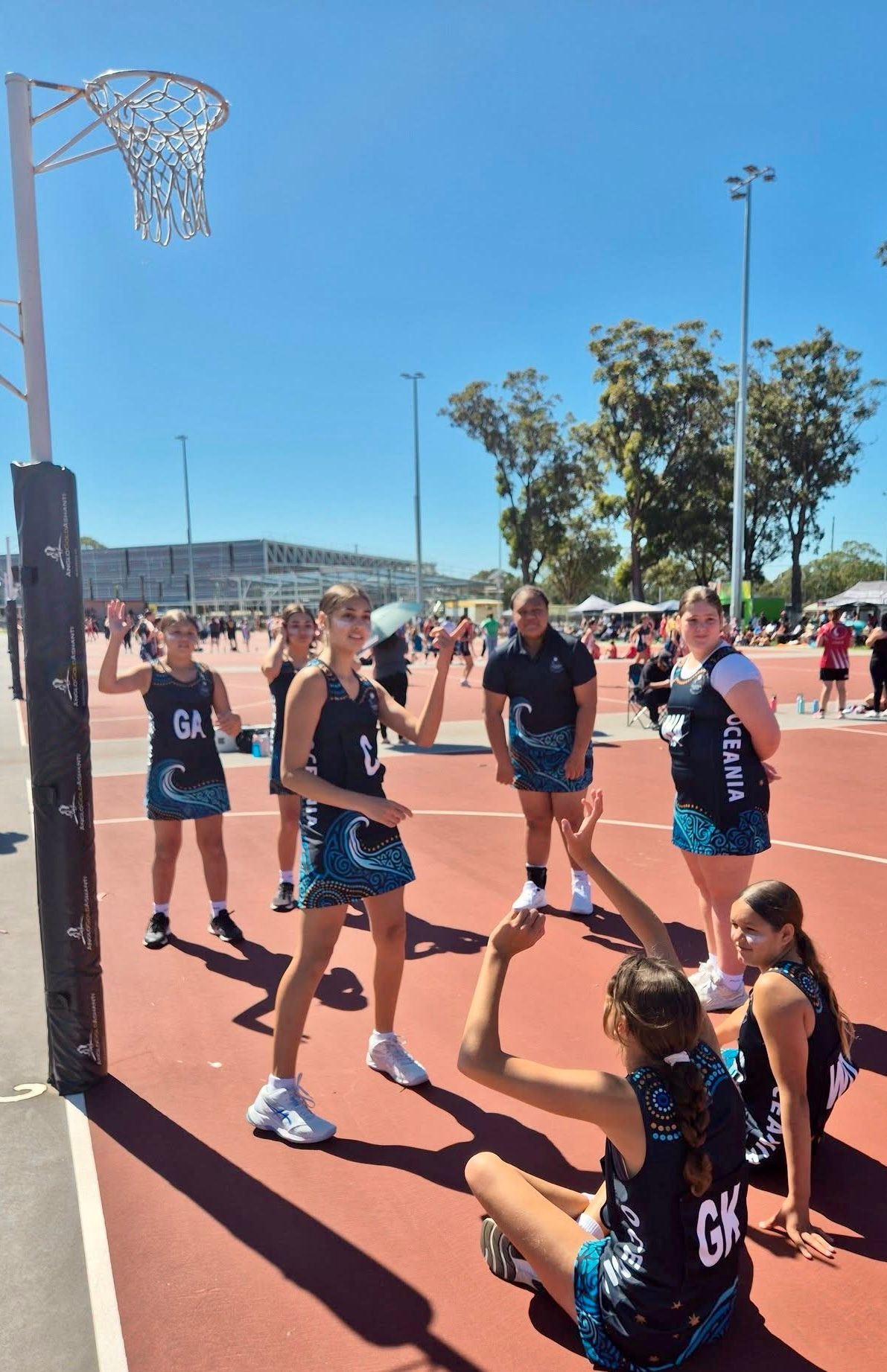 Netball players on a red court, under a blue sky, some in motion, others seated.
