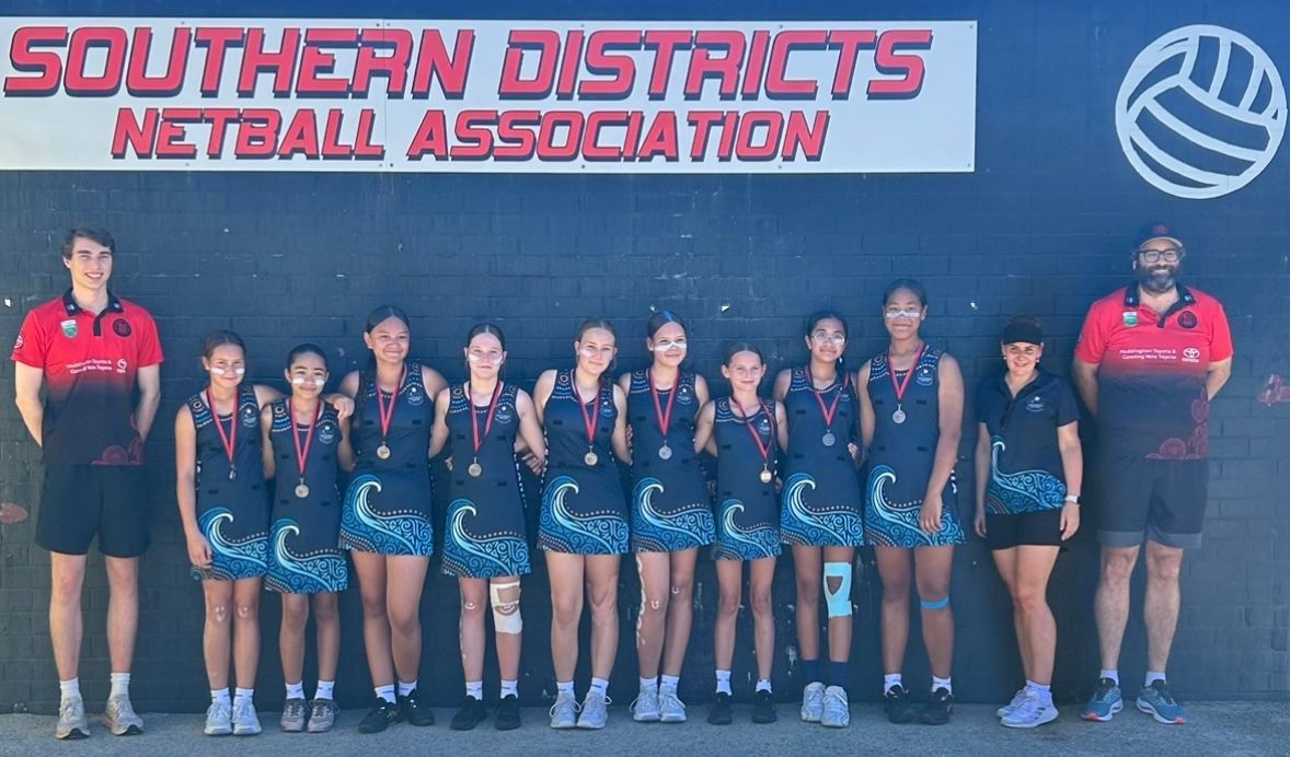 Netball team poses in front of a sign. They wear matching blue and white dresses, medals, and stand outside.