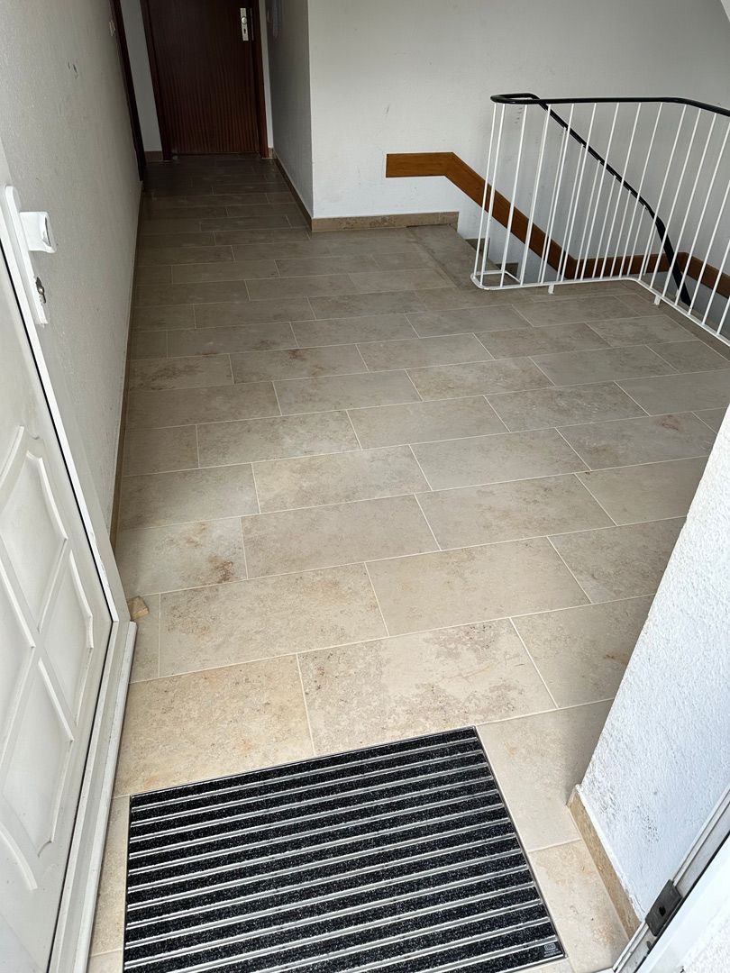 Eingangsbereich mit hellbraunem Fliesenboden, schwarzer Fußmatte, weißer Tür und Treppe mit weißem Geländer.