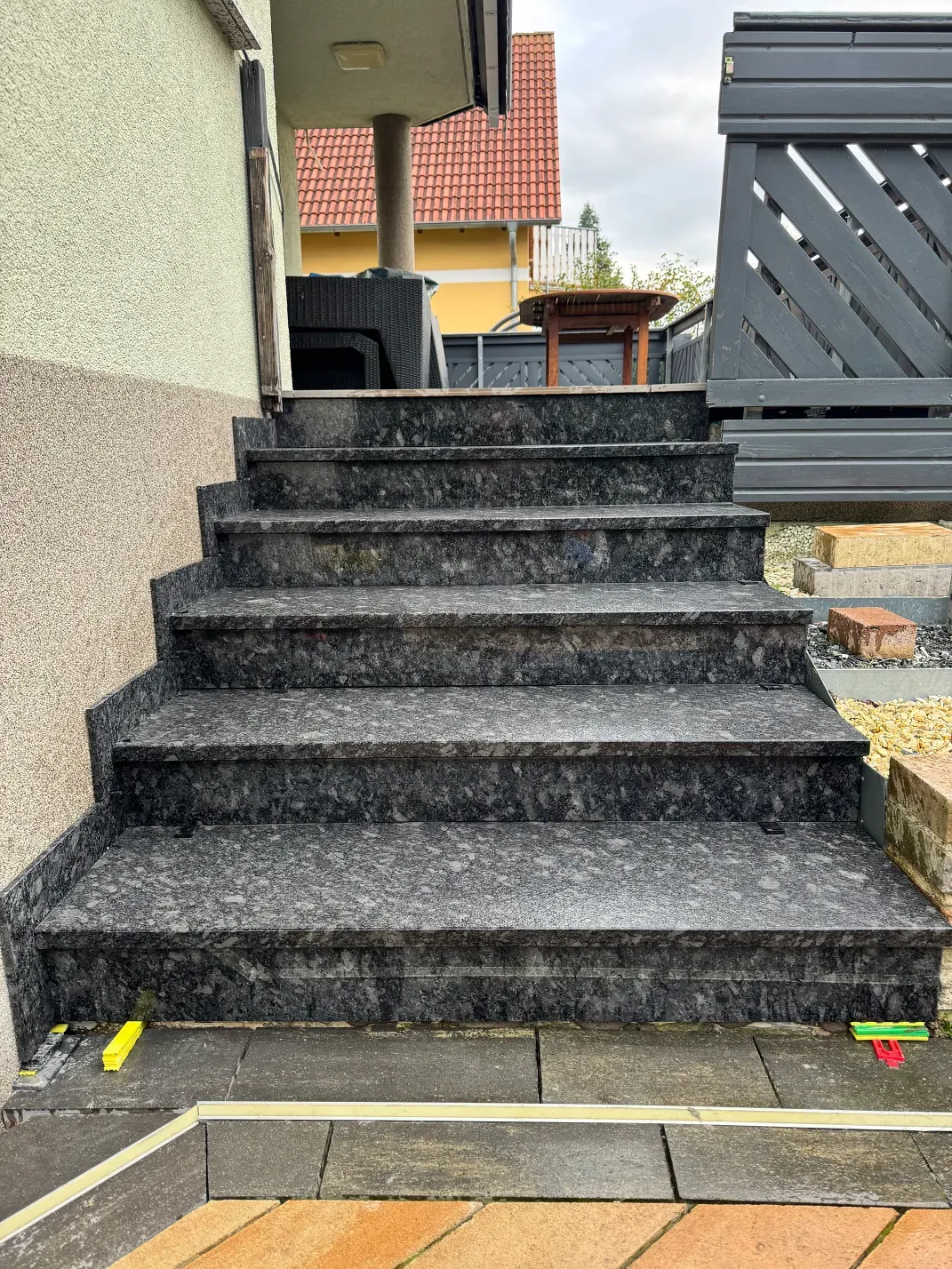 Granitstufen führen zu einem Gebäudeeingang; dunkelgraue Stufen, hellgraue Fassadenverkleidung und ein Holzzaun sind zu sehen.