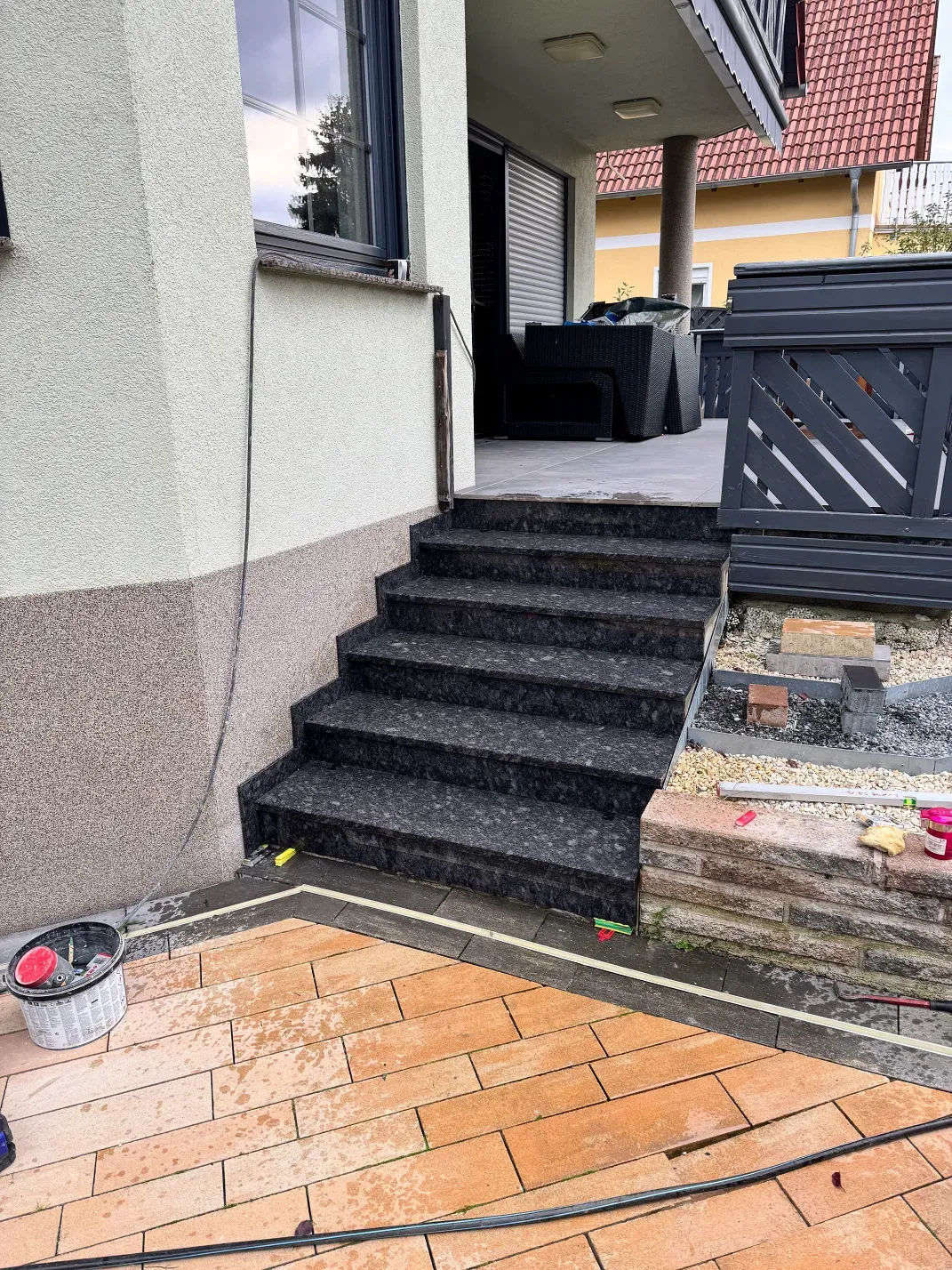 Außentreppe, die zum Eingang eines Gebäudes führt; dunkel gesprenkelte Stufen, helle Wände und Ziegelmauerwerk am Boden.