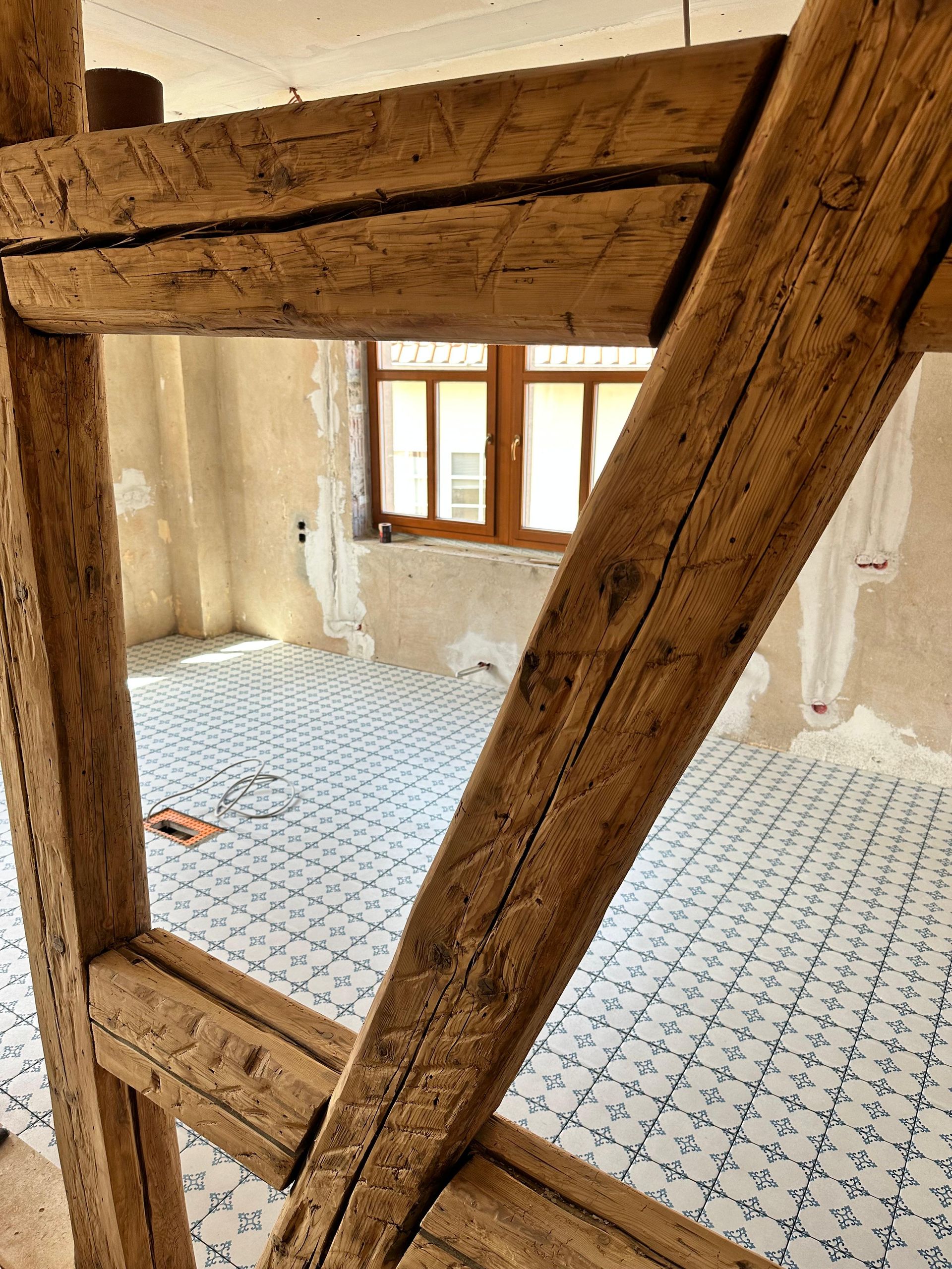 Holzbalken eines Gebäudes rahmen einen Raum mit Fenster, weißem Fliesenboden und sichtbarer Konstruktion ein.