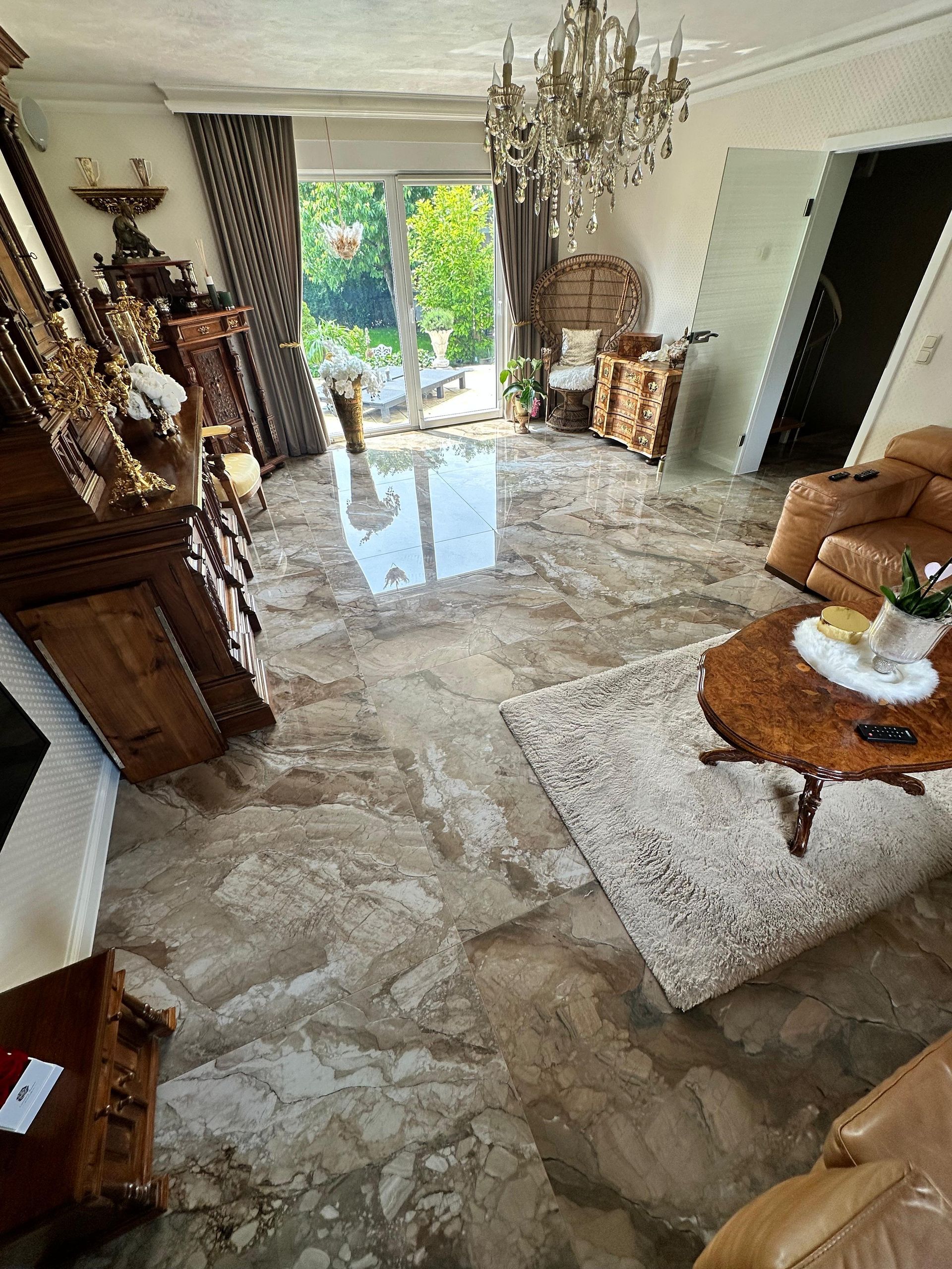 Wohnzimmer mit glänzendem Marmorboden, prunkvollen Möbeln und einem Kronleuchter. Sonnenlicht strömt durch eine Glastür.