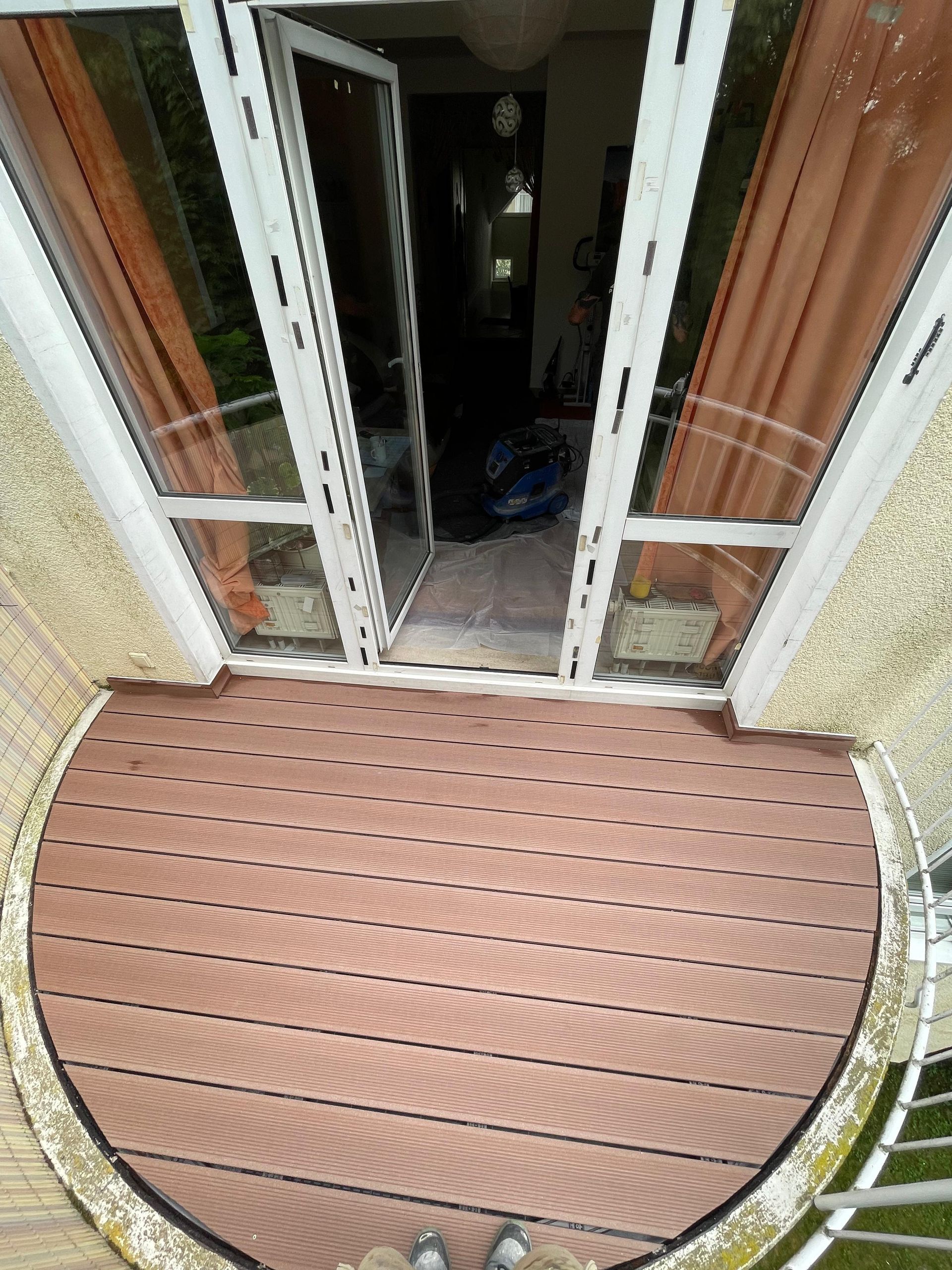 Ein Balkon mit braunen Dielen, offenen Glastüren und orangefarbenen Vorhängen.