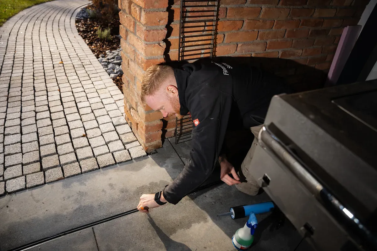 Ein Mann arbeitet auf Händen und Knien an einem Garagentoröffner in der Nähe einer Backsteinsäule und eines gepflasterten Weges.