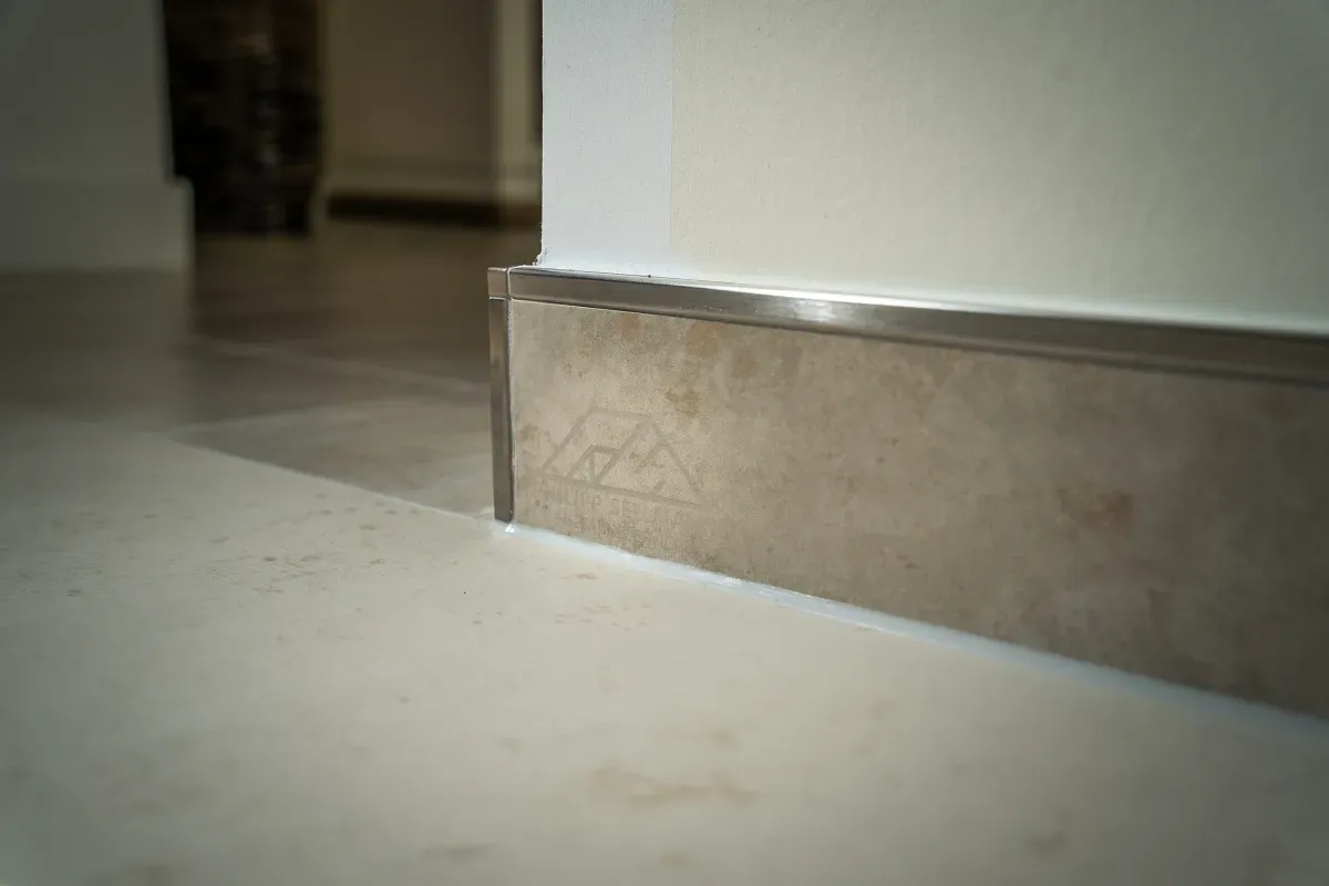 Nahaufnahme einer modernen Fußleiste. Eine Edelstahlkappe schließt eine helle, strukturierte Fußleiste vor einer weißen Wand und einem hellen Boden ab.