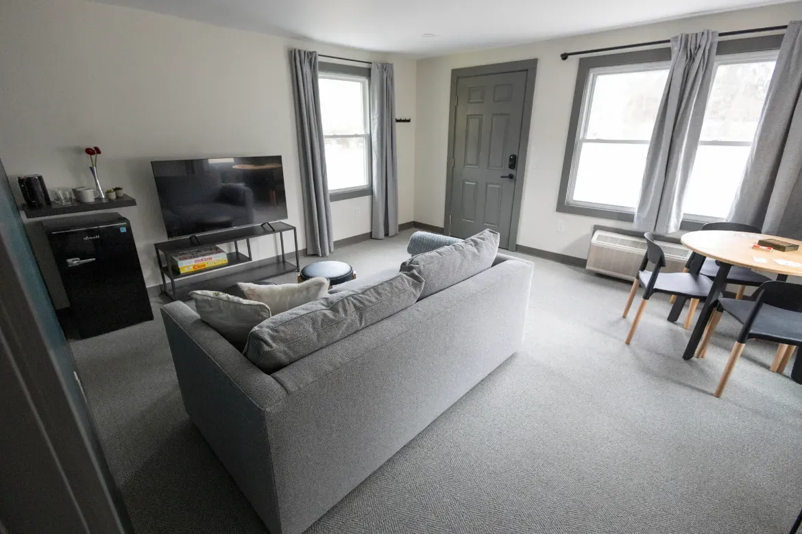 Bright living room with gray sofa, TV, windows, and small dining table near the door.