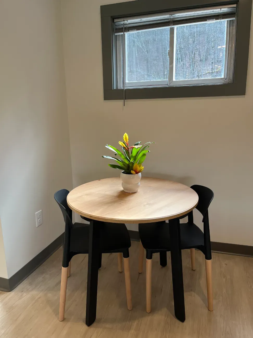 Small dining nook with a round wooden table, four black chairs, and a vase of flowers under a window.