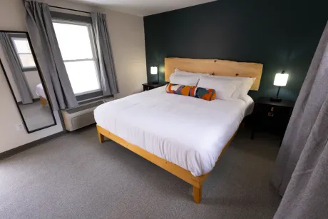 Bright bedroom with a wooden bed, white linens, dark green accent wall, and two windows.
