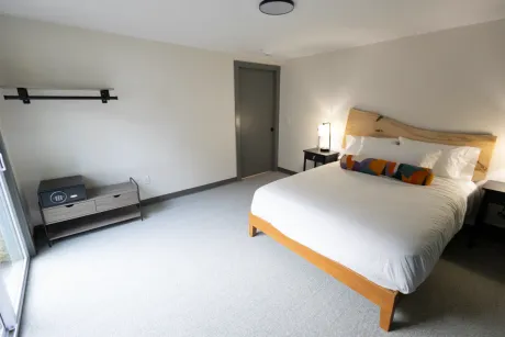 Minimal bedroom with a bed, bench, nightstand, and door on a light carpeted floor.