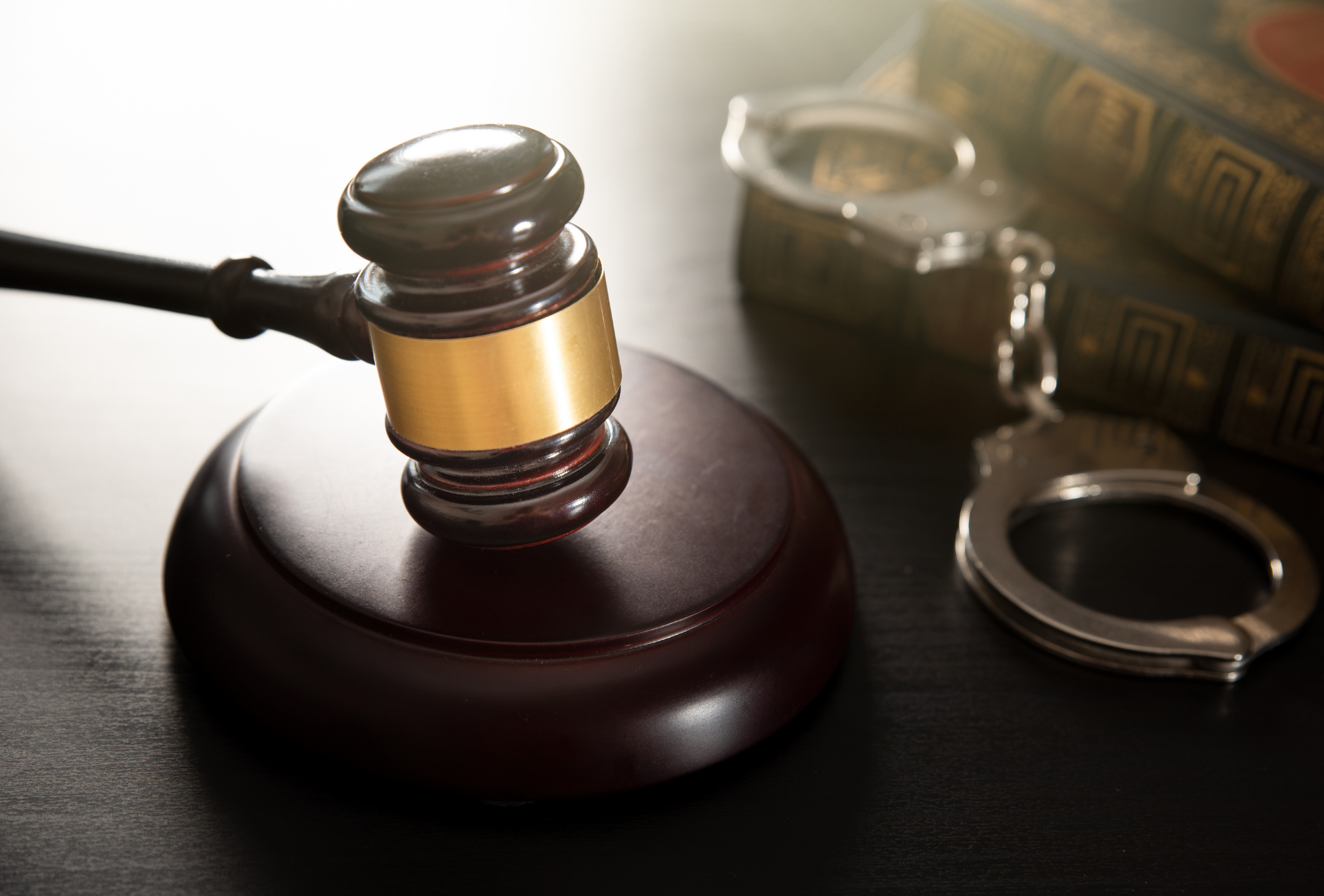 Gavel on block next to handcuffs and law books on a table, representing legal proceedings.