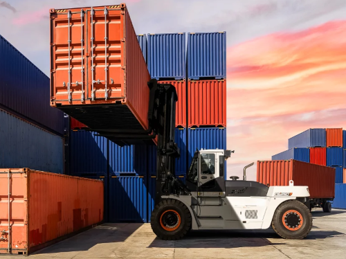 Forklift lifting a red shipping container in a busy port with stacked containers, under an orange and blue sky.