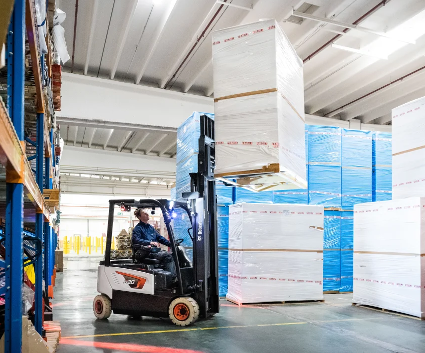 Forklift in warehouse lifting a large wrapped package, with stacks of goods in the background.