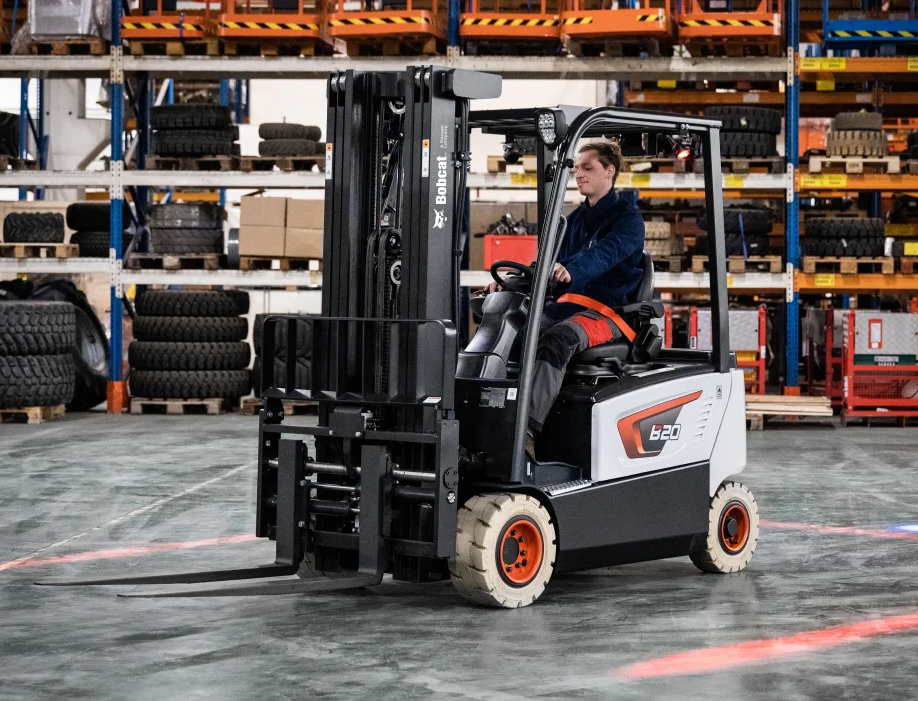 Man operating a white and black forklift in a warehouse. Red safety light visible on floor.