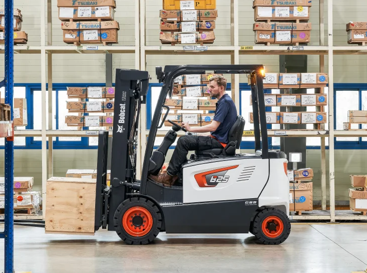 Man operating a Bobcat forklift in a warehouse, transporting a wooden crate.