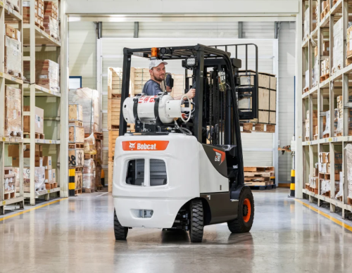 A man operates a white Bobcat forklift in a warehouse with shelving full of boxes.