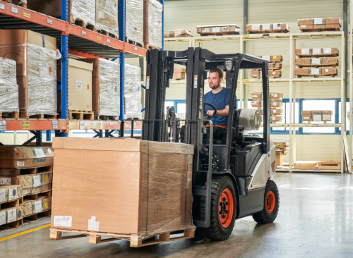 A man operates a white Bobcat forklift in a warehouse with shelving full of boxes.