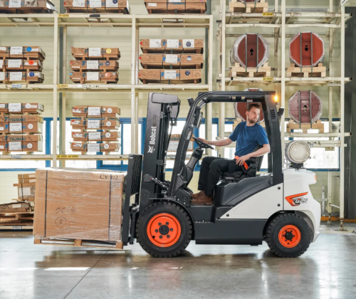 A man operating a Bobcat forklift inside a warehouse, moving a wooden crate.