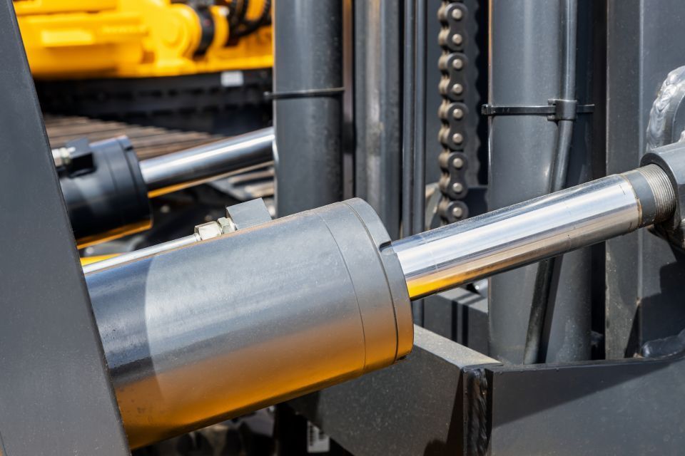 Close-up of a forklift's hydraulic cylinder, metallic and black, with a yellow machine visible in the background.