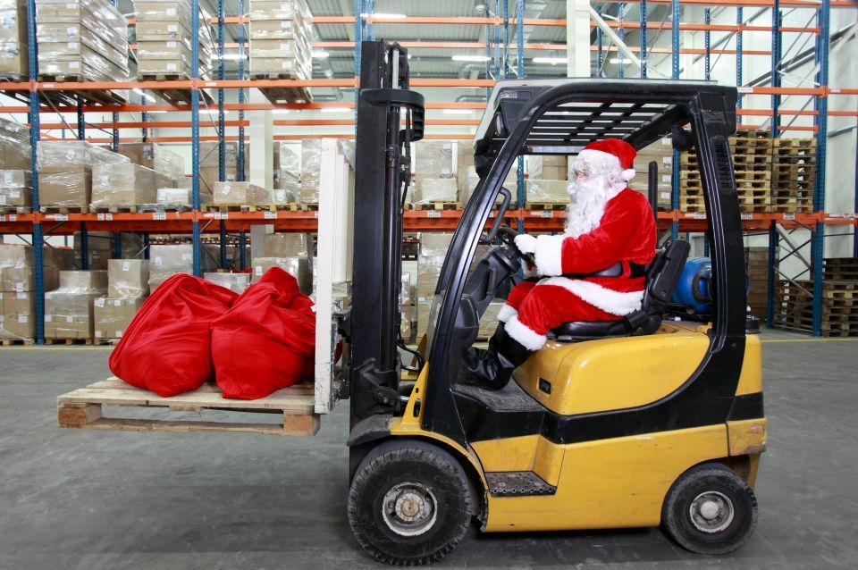 Santa Claus driving a forklift in a warehouse, carrying red gift sacks on a pallet.