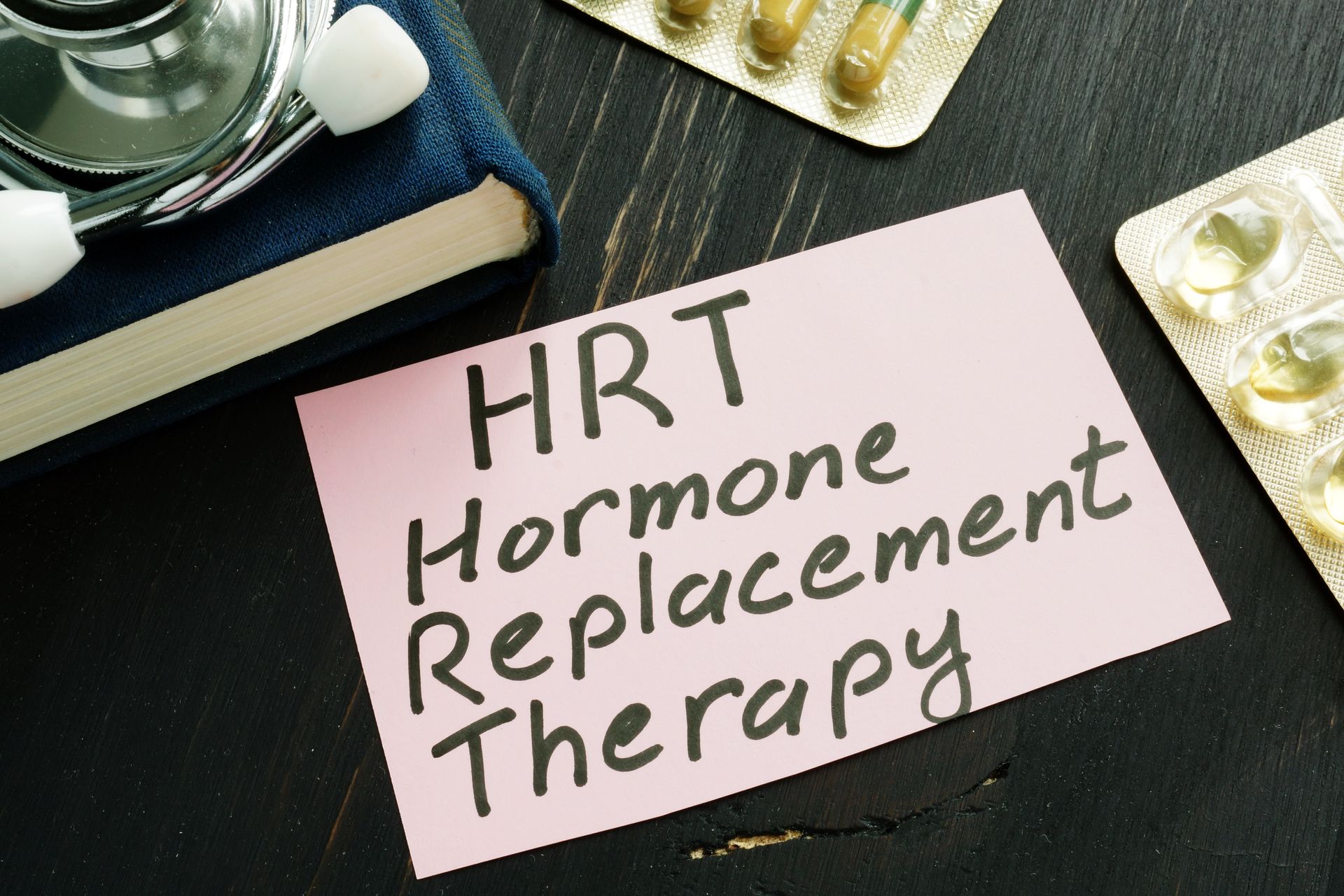 Pink note reading “HRT Hormone Replacement Therapy” on a dark desk with pills and medical items nearby Pink note reading “HRT Hormone Replacement Therapy” on a dark desk with pills and medical items nearby