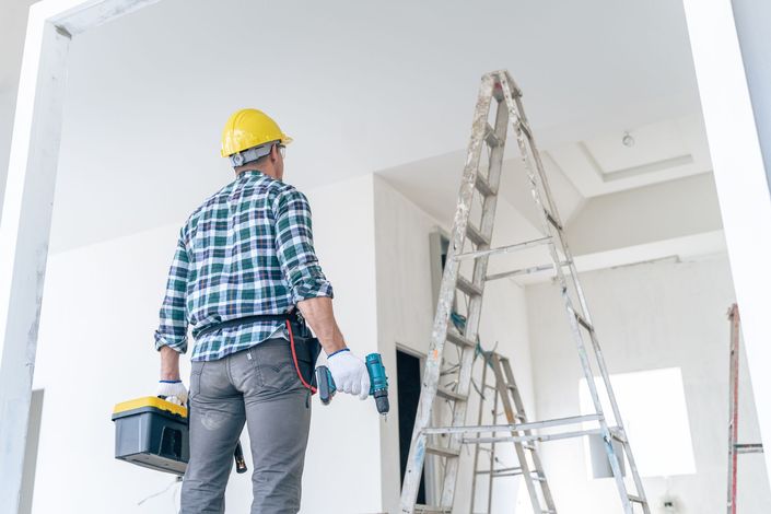 Operaio edile con casco giallo, cassetta degli attrezzi e trapano, che guarda verso il soffitto vicino a una scala.