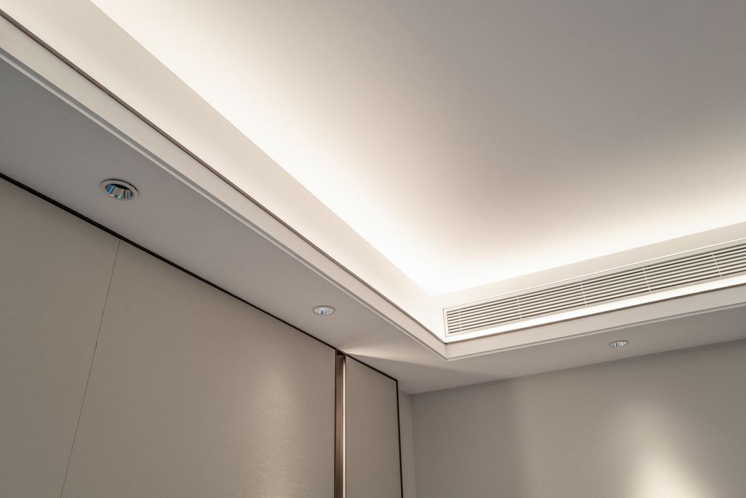 Un soffitto angolare con illuminazione da incasso, una parete beige chiaro e un piccolo allarme antincendio.