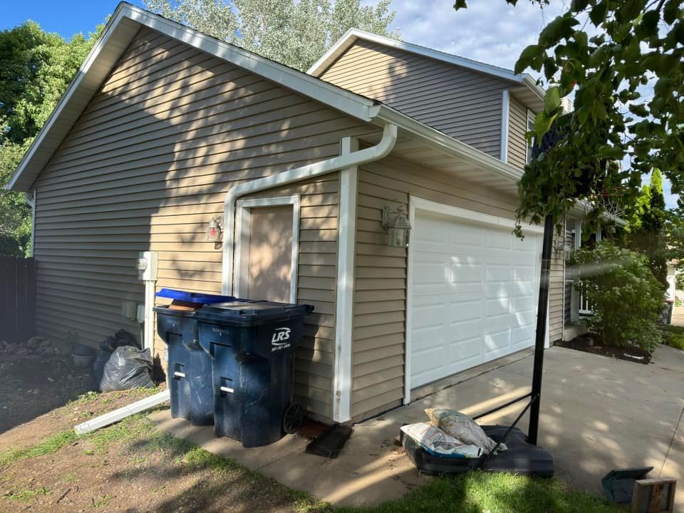 Tan and brown two-story house with a white garage door, trash bins, and a basketball hoop.