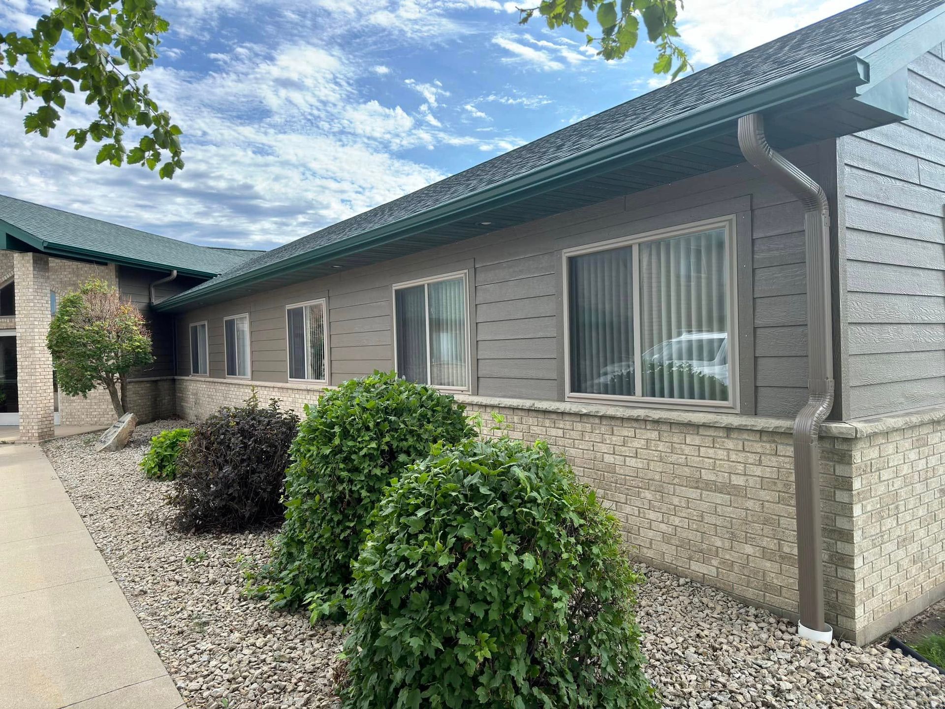 Building with green roof, tan siding, and stone base; bushes and gravel in front.