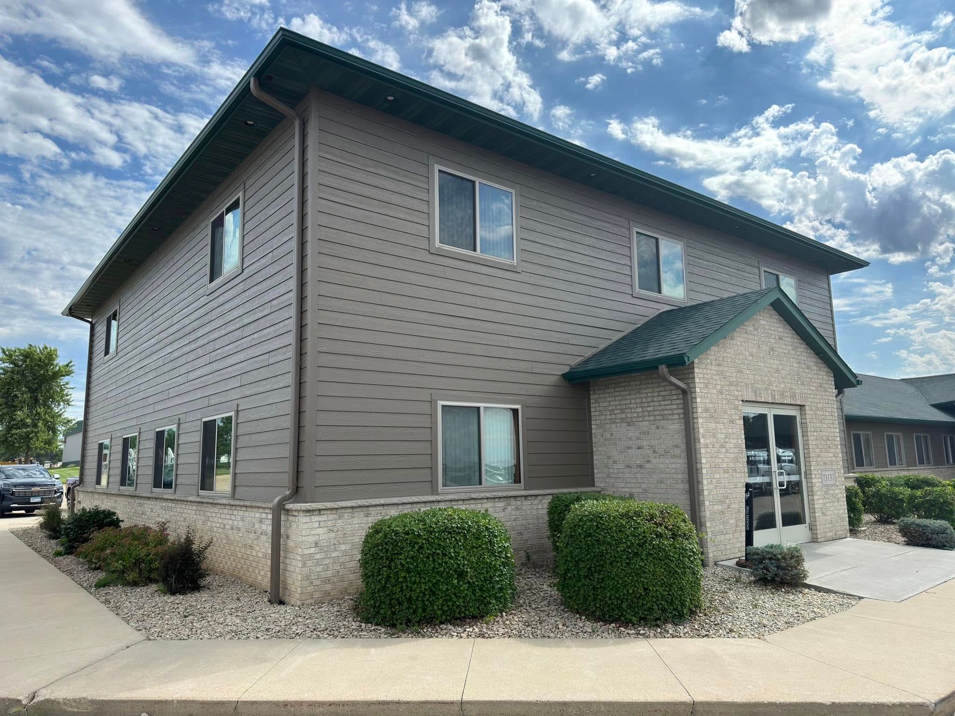 Two-story building with tan siding, green roof, and small bushes. Entrance has a brick facade.