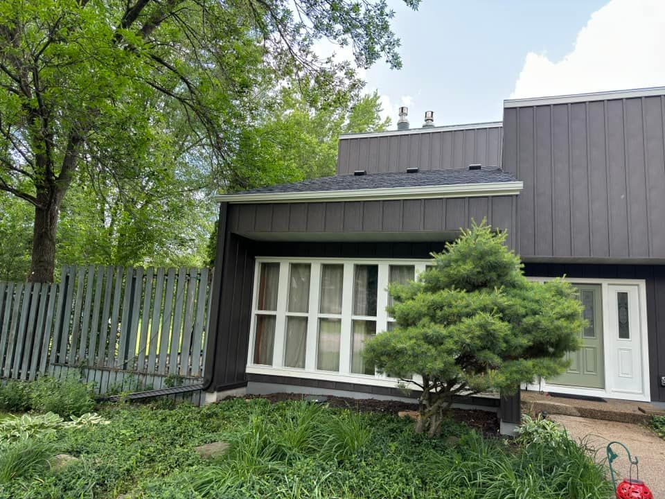 Gray house with white-framed windows, a small evergreen tree, and a weathered wooden fence.