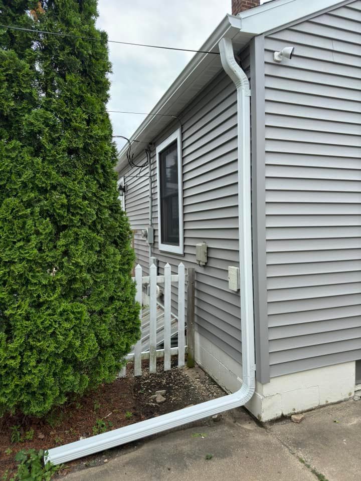 White gutter on a gray house with a small white picket fence and evergreen shrub in a residential setting.