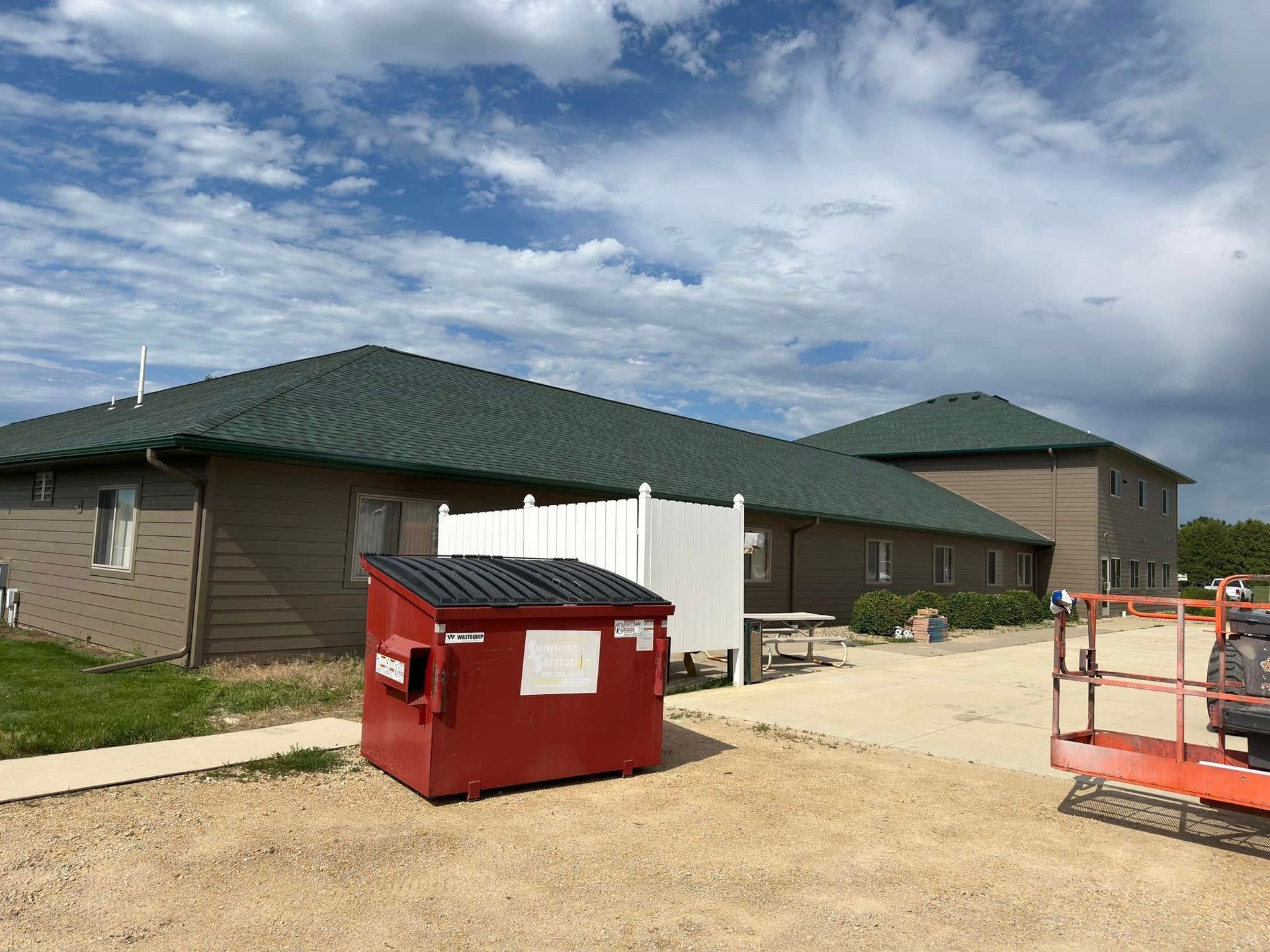 Brown building with green roof, red dumpster, white fence, gravel lot, cloudy sky.