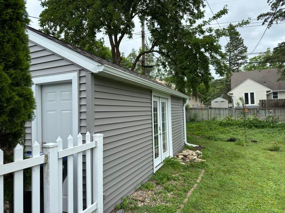 Gray shed with white trim, door, and French doors. White picket fence in front. Green grass and trees.