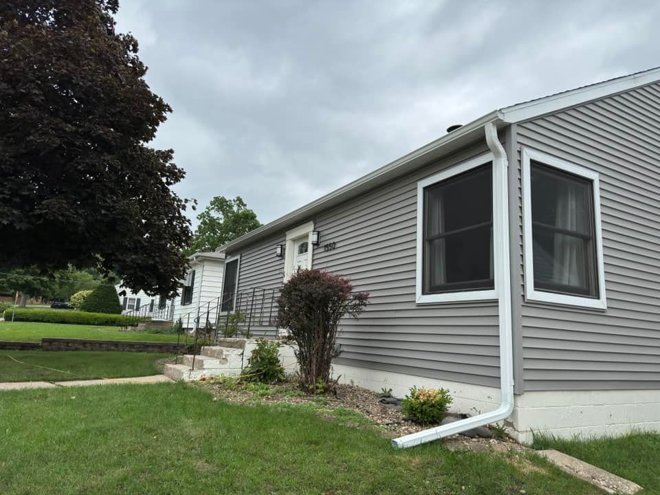 Gray-sided house with white trim, two windows, and a downspout under an overcast sky.