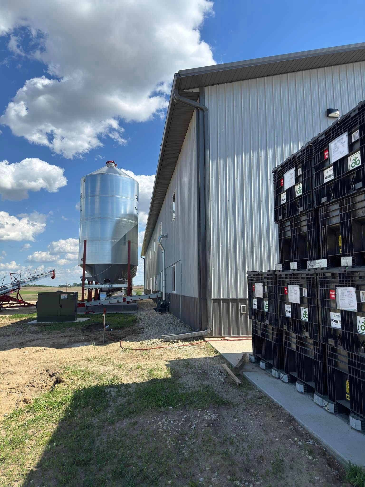 A tall, corrugated metal building with a silo and stacked black containers, under a cloudy blue sky.