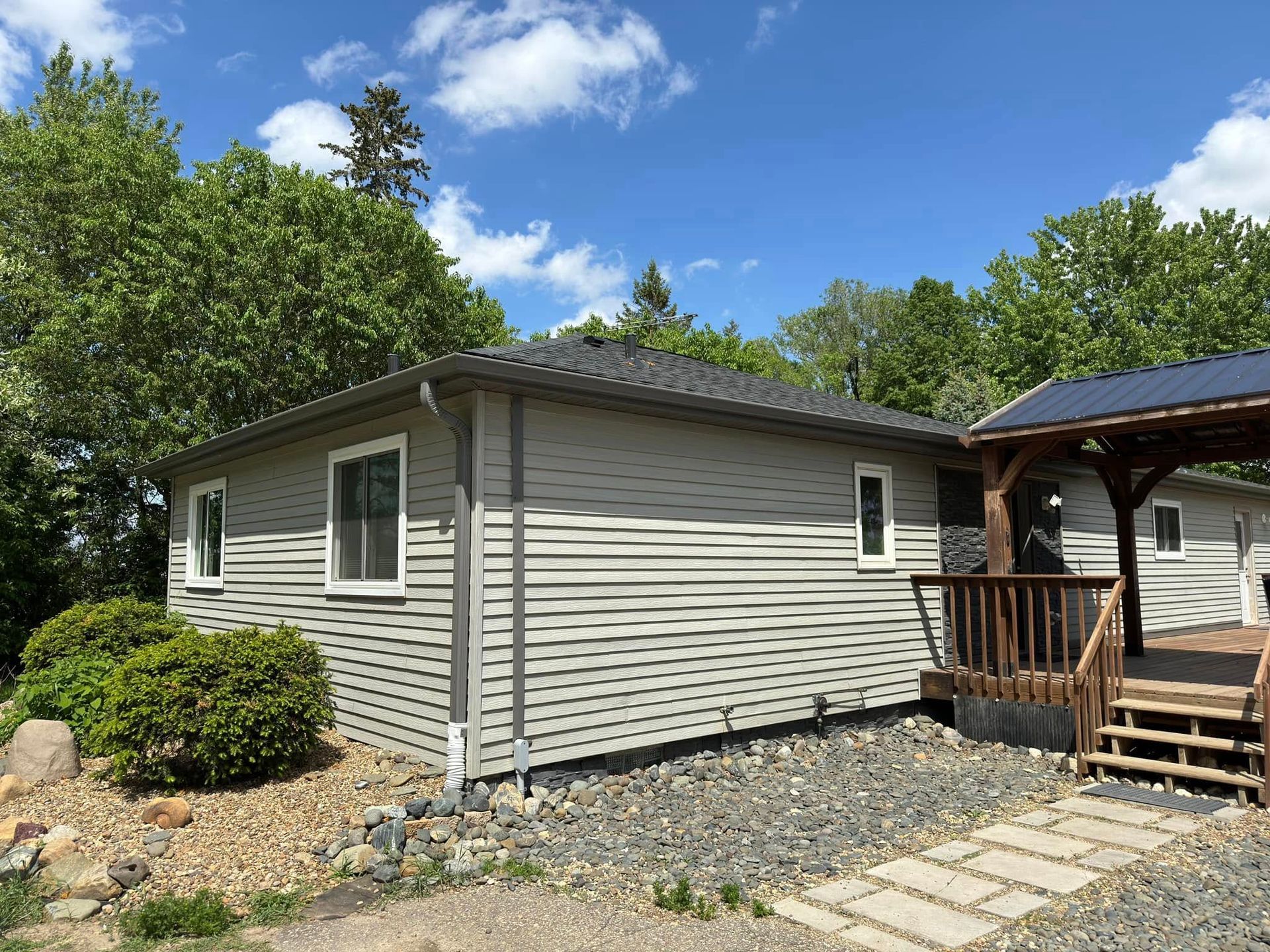 A light gray house with a dark gray roof sits on a gravel yard next to a wooden deck under a blue roof.