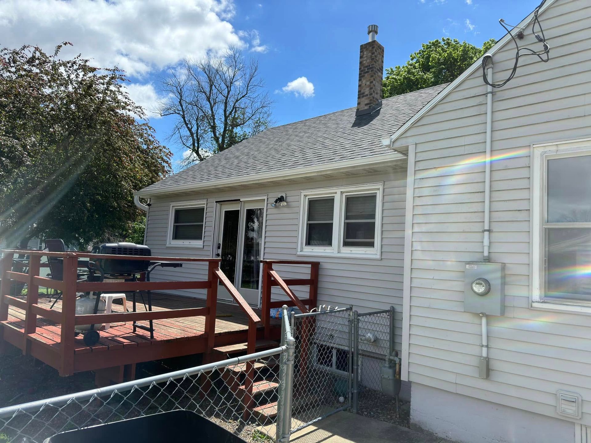 Backyard view of a gray house with a red deck and a chain-link fence on a sunny day.