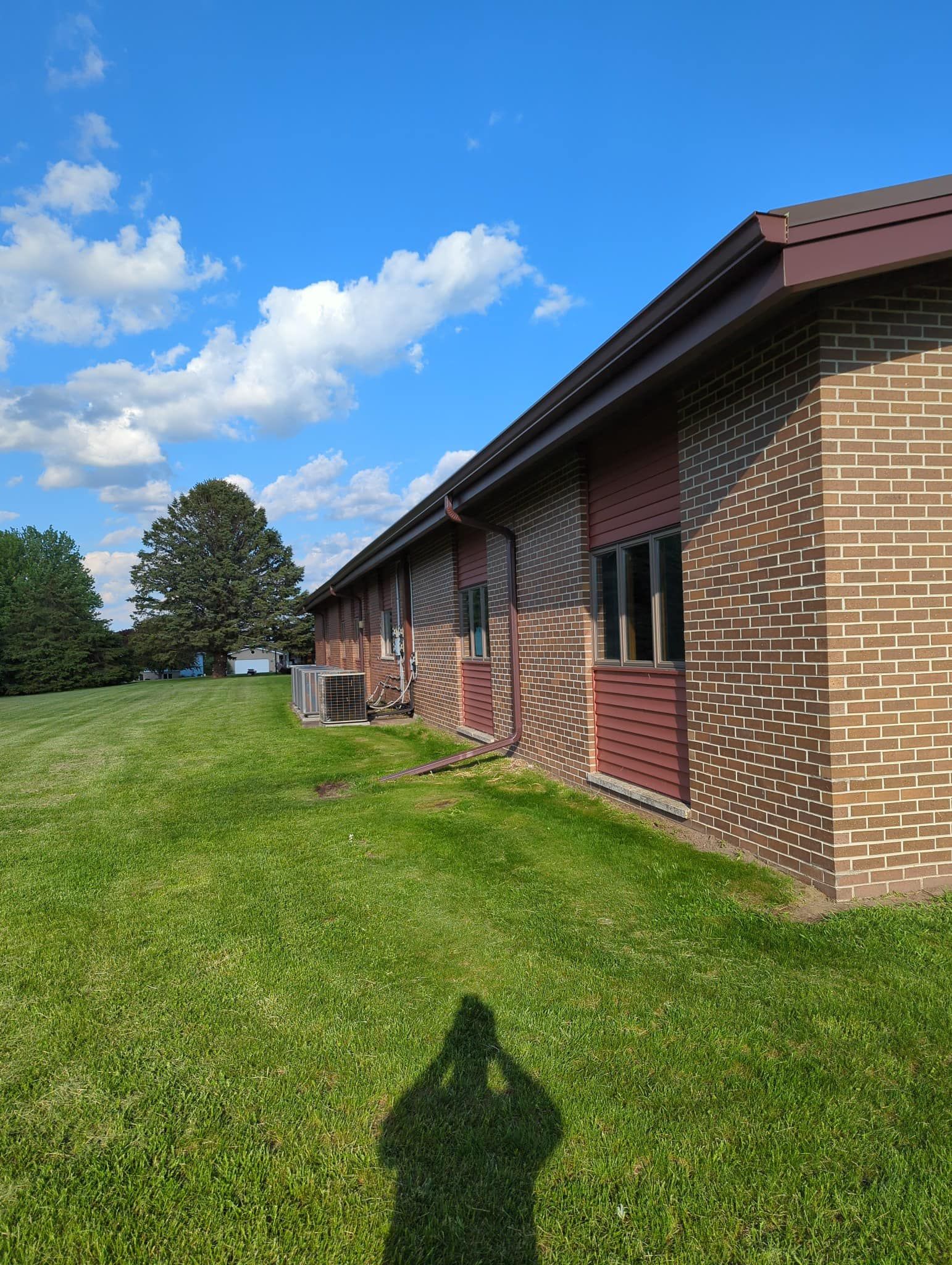 Brick building with brown roof, green lawn, blue sky with clouds. Shadow in foreground.