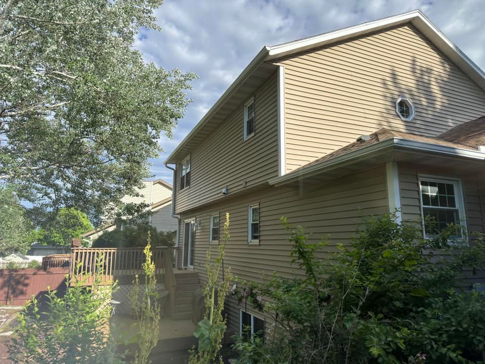 Tan house with siding and a wooden deck. Lush green trees and sky are visible.