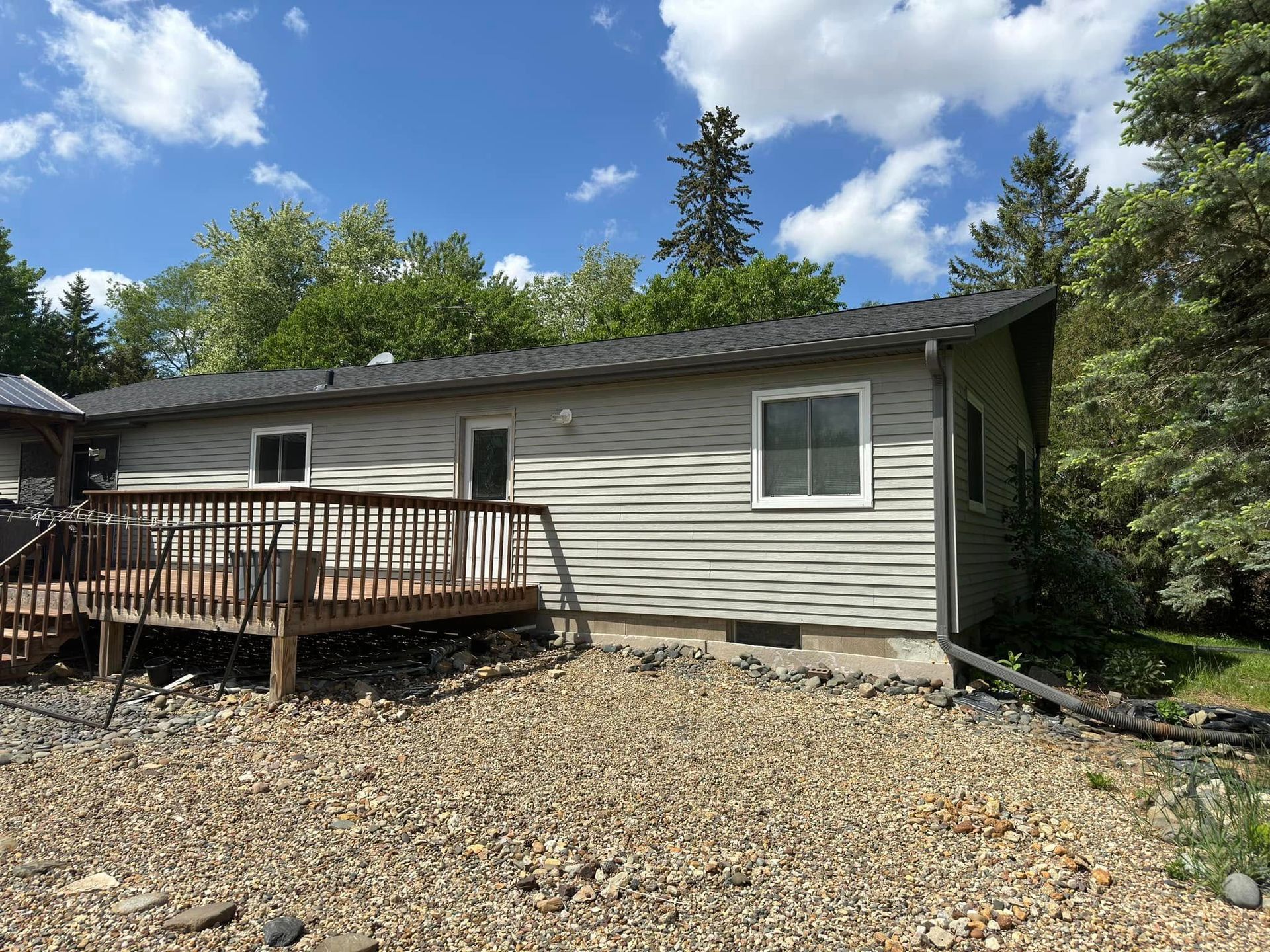 House with light siding, dark roof, wooden deck, and gravel yard. Cloudy sky, trees.