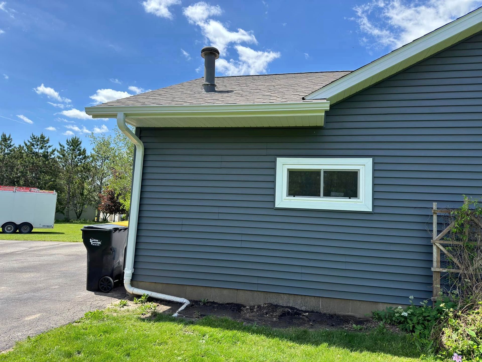 Blue-sided house with white trim, window, and gutters; a black trash can sits on the driveway.