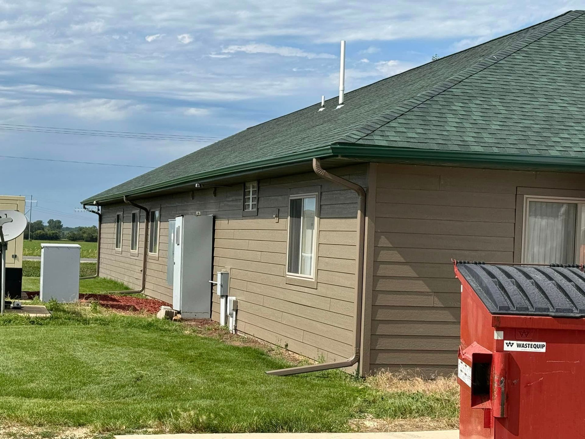 Tan building with green roof, a white door, and red dumpster in yard.