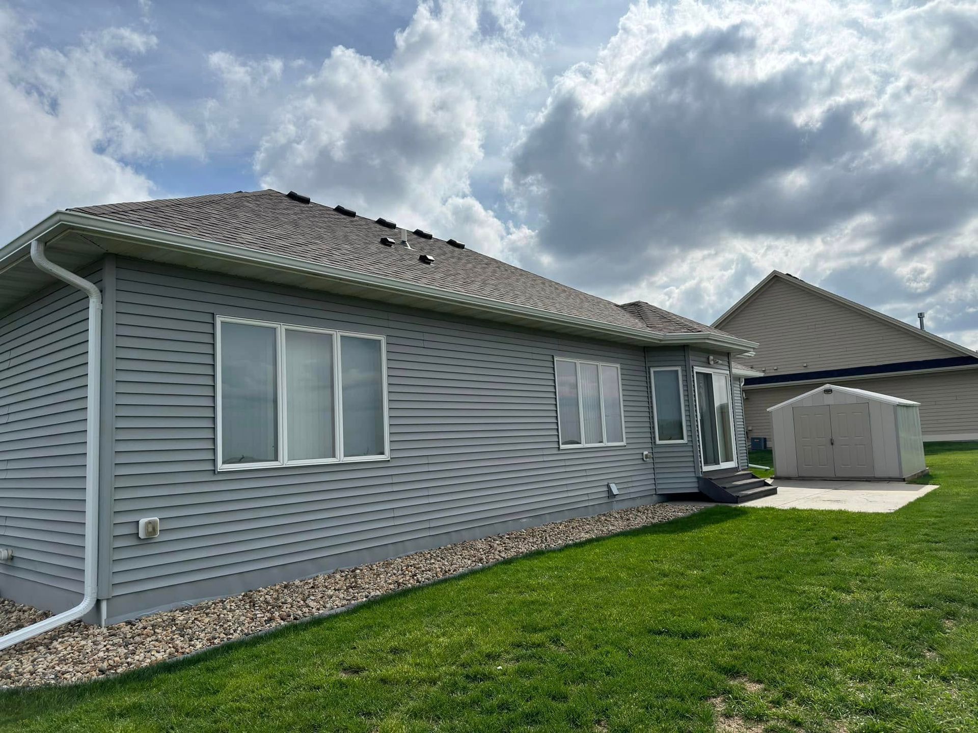 Gray-sided house with white trim, windows, and roof against a cloudy sky. Lawn in foreground, shed in the background.