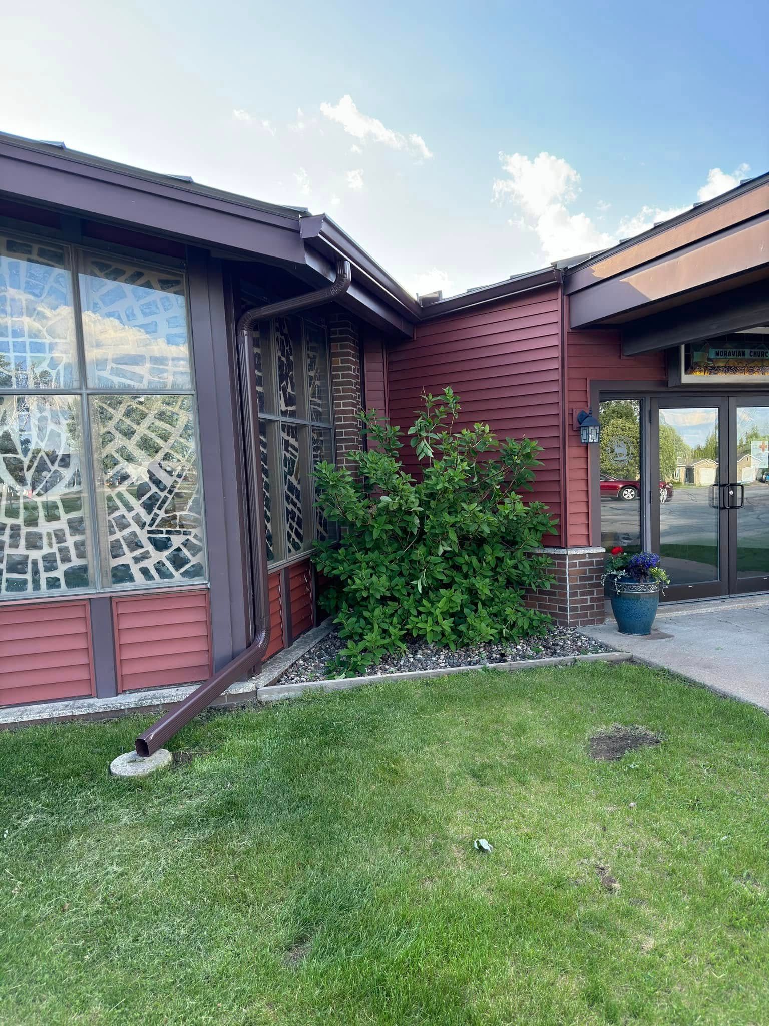 Red building with large stained-glass window, green lawn, bush, and glass entrance door.