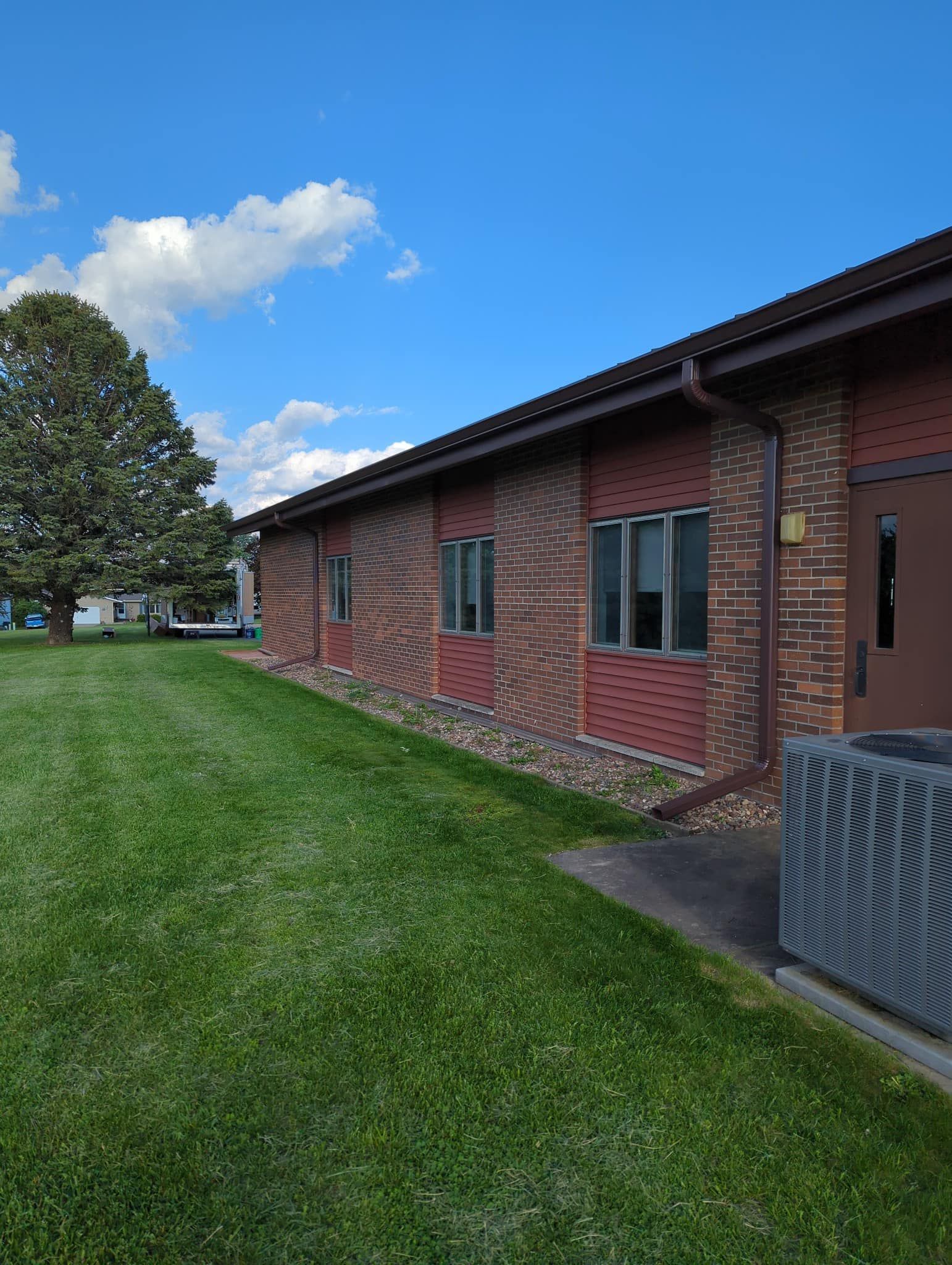 Brick building with windows and a dark brown door, green lawn and tree, blue sky.